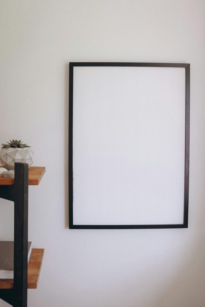 A clean, minimalist blank frame on a neutral-colored wall with a decorative shelf nearby.