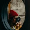 A woman in a red beanie photographs indoors with a vintage camera.